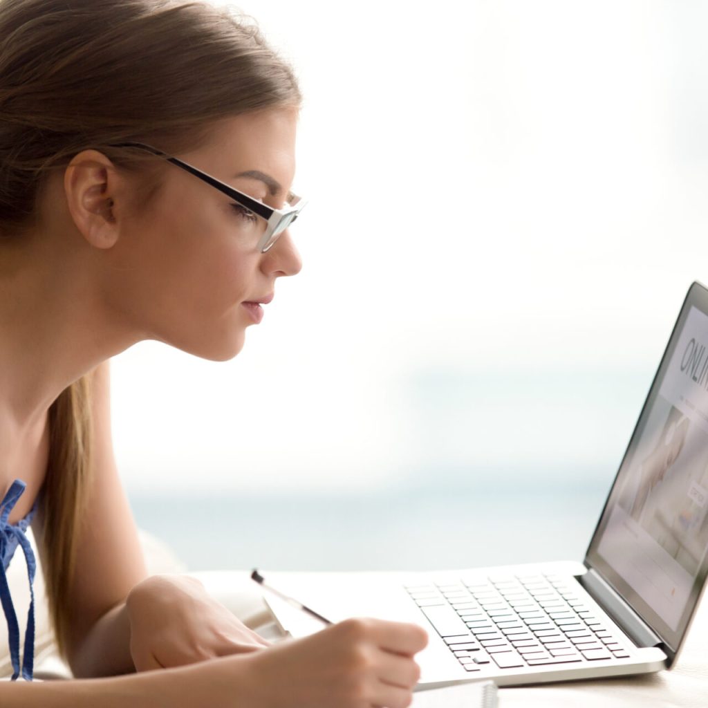 Female student studying online with laptop Female student in eyeglasses writing in notebook while visiting online study courses web page in Internet. Millennial woman distantly learning with laptop at home, getting new knowledge or profession